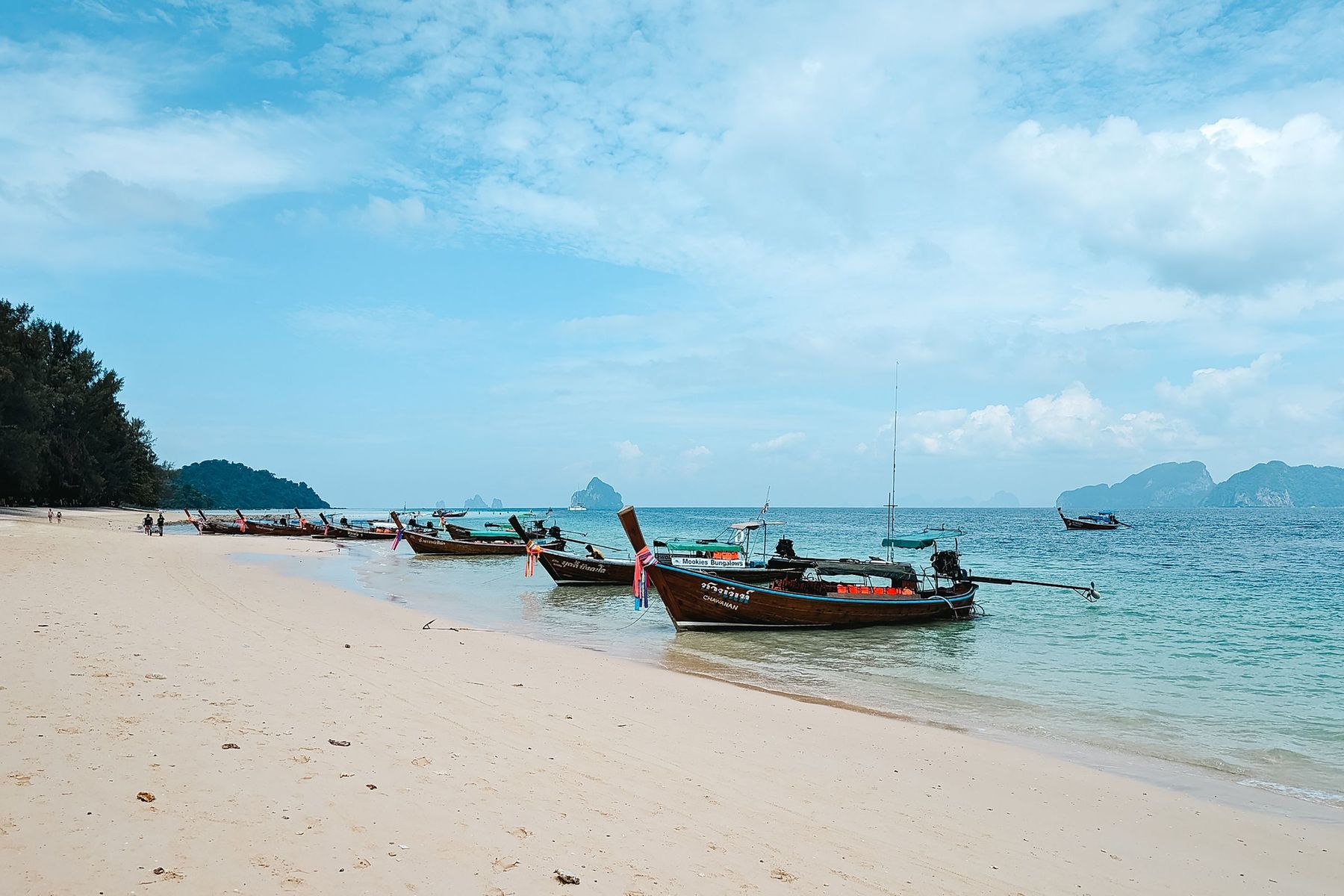 Longtail boats moored off a quiet white sand beach with islands on the horizon