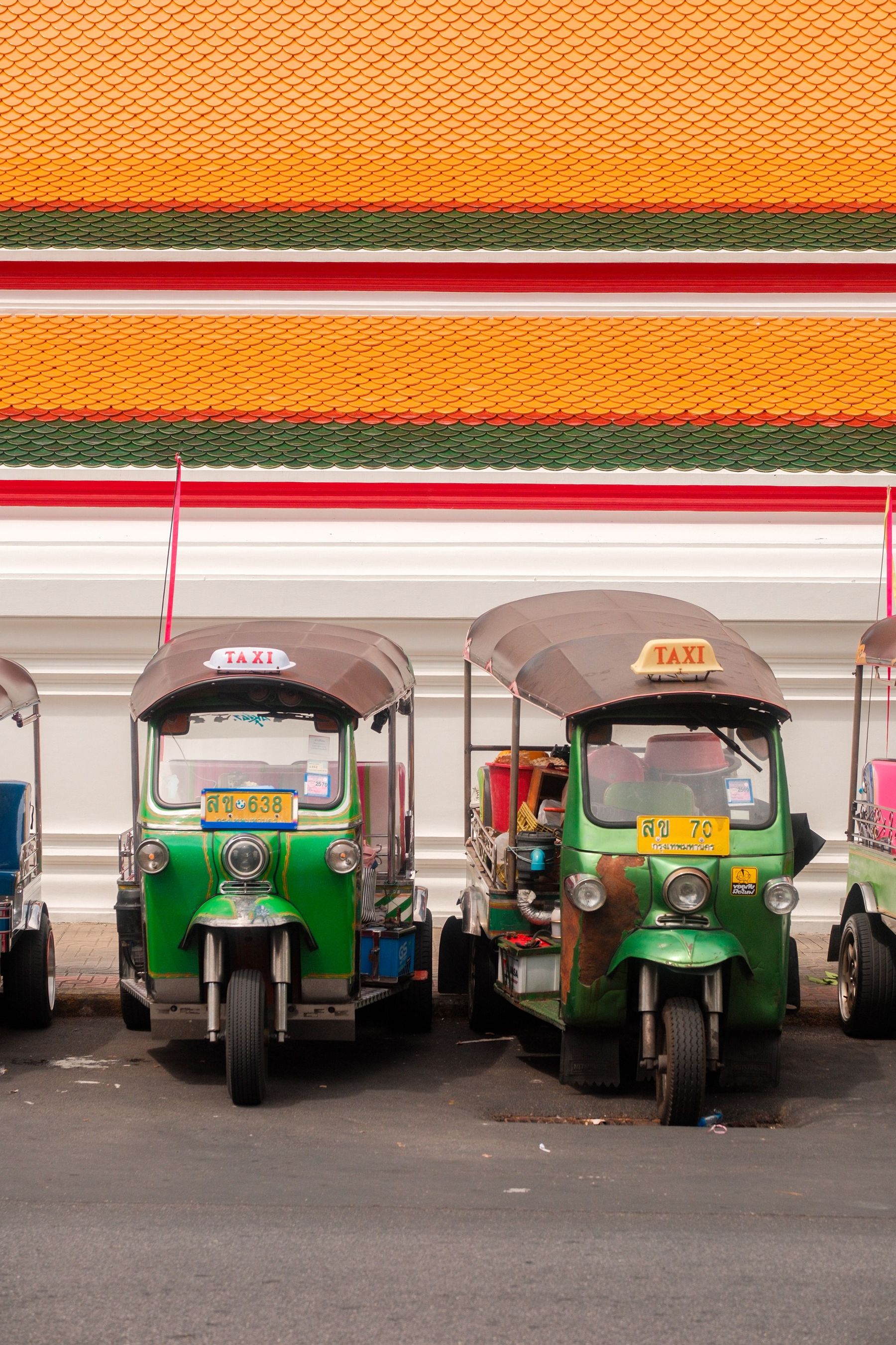 Two green and yellow tuk-tuks parked in front of a temple's orange and white wall